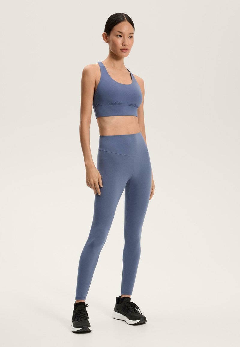 Világoskék sportmelltartó és hozzá illő, magas derekú leggings rugalmas anyagból, fekete sportcipővel párosítva. Sima textura, minimális dizájn.