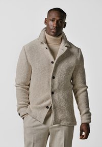 Beige gewobene Jacke mit einem Kragen, ausgestattet mit schwarzen Knöpfen und einer strukturierten Oberfläche. Getragen über einem cremefarbenen Rollkragenpullover, kombiniert mit hellen Hosen.
