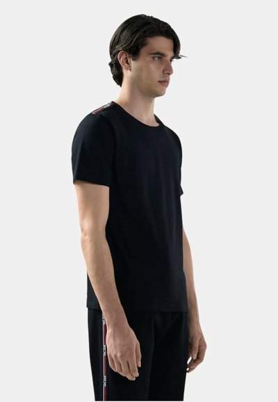 Giovane uomo con maglietta nera e pantaloni neri con dettagli a strisce rosse e bianche, in piedi di profilo verso destra su uno sfondo chiaro e uniforme.