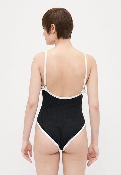 Maillot de bain noir avec accents blancs, vue arrière. Présente des bretelles fines, un dos décolleté, et un matériau texturé avec un léger éclat.