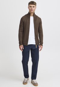 Cardigan marrone a maglia con dettagli a costine, colletto zip, indossato sopra una camicia bianca, abbinato a pantaloni blu scuro e sneaker grigio-marroni.