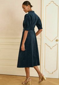 Abito midi in velluto a coste blu navy con maniche a pompon, vita cinturata e sandali con tacco a blocco dorati. Texture morbida e silhouette semplice.