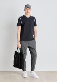 Sort T-shirt med hvide detaljer, grå bukser og en grå kasket. Modellen holder en sort tote bag og har hvide sneakers på med struktureret sål.