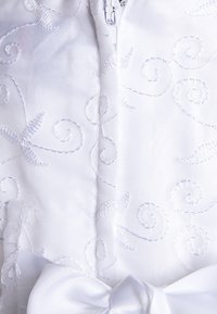 Valkoinen kangas, jossa on brodeerattuja kiehkuja. Varustettu vetskalla ja suurella satiinikaarella alhaalla, luoden tekstuurista ja eleganttia ilmettä.