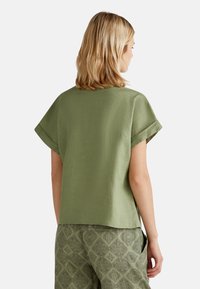 Blusa verde oliva con un taglio squadrato, maniche corte con risvolto e un tessuto dalla trama liscia, abbinata a pantaloni verdi con motivo.