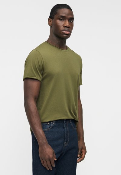 Άνδρας με κοντομάνικο μπλουζάκι χακί και σκούρο μπλε τζιν, γυρισμένος ελαφρώς πλάγια μπροστά σε απλό φωτεινό φόντο.