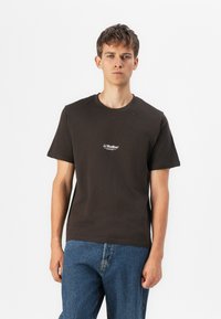 Camiseta de algodón marrón oscuro con mangas cortas, cuello redondo y pequeño logo blanco de 'JJ Studios' en el pecho. Combinada con jeans de mezclilla azul claro.