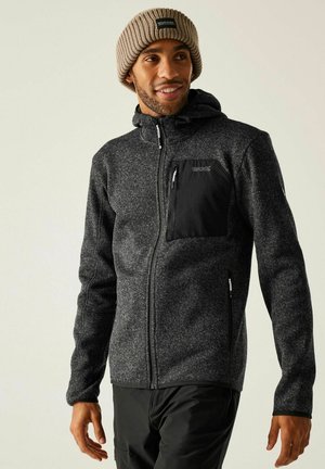 Hombre con chaqueta gris texturizada con cremallera y bolsillo negro en el pecho, y gorro de punto marrón, sonriendo y mirando hacia la derecha.