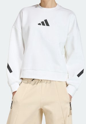 Osoba nosząca białą bluzę z czarnym trójkątnym logo na klatce piersiowej i ramionach, w zestawieniu z beżowymi spodniami z kieszeniami na zamek.