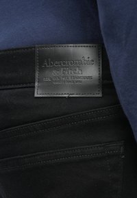 Črn usnjen znak z vgraviranim napisom "Abercrombie & Fitch" na črnem denim pasu, v kombinaciji s temno modrim materialom nad njim.