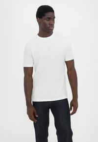 Hvit t-skjorte med rund hals og tettsittende design. Har forhøyet logo tekst på brystet, kombinert med mørke denimjeans. Glatt bomullstekstur.