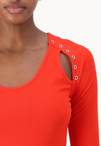 Rotes Langarmoberteil mit Rundhalsausschnitt und ausgeschnittenen Schultern. Verfügt über silberne Ösen entlang der Schultern für zusätzliche Details und Textur.