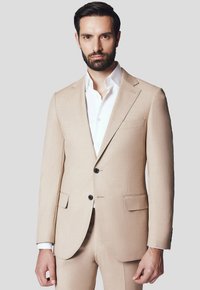 Beżowa marynarka w garniturze z dwoma guzikami, klapami w szpic i bocznymi kieszeniami. Nosi się ją na białej koszuli. Gładka tekstura.