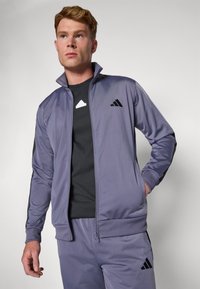 Lila sportos tréningdzseki, teljes cipzárral, álló gallérral és fekete részletekkel. A mellkason három fekete Adidas csík található.