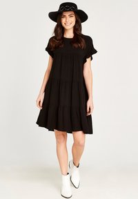 Schwarzes, gestuftes Kleid mit kurzen Ärmeln, hergestellt aus weichem Stoff. Begleitet von einem schwarzen Hut und weißen Knöchelstiefeln. Lässig und locker geschnitten.