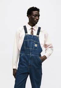 Άνδρας που φορά μπλε τζιν σαλόπετα πάνω από ένα ανοιχτό ροζ πουκάμισο με καφέ γραβατάκι με σχέδιο, στέκεται με το ένα χέρι στην τσέπη, κοιτώντας κάτω.