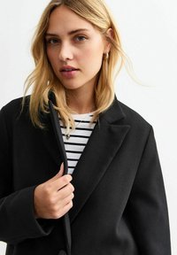 Manteau noir en mélange de laine au design ajusté, avec un grand col et un bouton de fermeture à l'avant, porté par-dessus une chemise rayée blanche et noire.