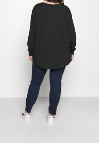 Svart långärmad topp med rundad nederkant, kombinerad med mörkblå jeans och vita sneakers med svarta detaljer. Slät tygtextur.