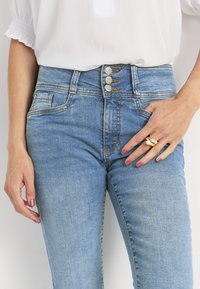 Korkeavyötäröiset, vaaleansiniset farkut, joissa on viiden napin sulku, kontrastiväriset saumat ja sivutaskut, yhdistetty valkoiseen paitaan.