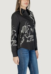 Camicia nera con design di testo astratto bianco, collo a bottoni, maniche lunghe e orlo leggermente curvo, abbinata a jeans blu.