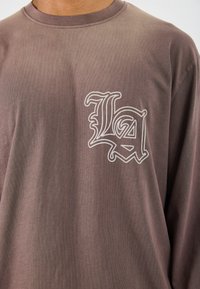 Pikem varrukaga särk muted pruunist kangast, millel on sile tekstuur. Esiküljel vasakul rinnal on valge broderi "LA" logo.