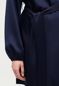 Κοντινή λήψη ενός ατόμου που φοράει φόρεμα από ναυτικό σατέν με δεμένη ζώνη στη μέση και μακριά μανίκια με ελαστικά τελειώματα.