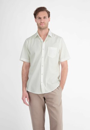 Mandlig model står med den ene hånd i lommen iført lyse, kortærmede, stribede skjorte og beige bukser mod en ensfarvet baggrund.