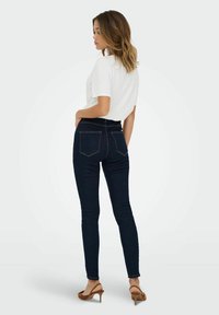 Donkerblauwe skinny jeans met een hoge taille, gecombineerd met een witte shirt met korte mouwen en bruine haksandalen, met achterzakken.