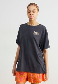Svart bomull T-skjorte med rund hals og korte ermer, med en liten, kontrastfarget Roxy-logo på brystet; parret med lyse, mønstrede shorts.