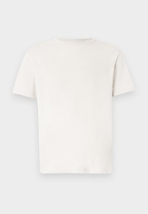 Ung mann iført hvit t-skjorte, beige rutete bukser, svarte slip-on sko og en liten svart skulderveske, stående mot en ensfarget bakgrunn.