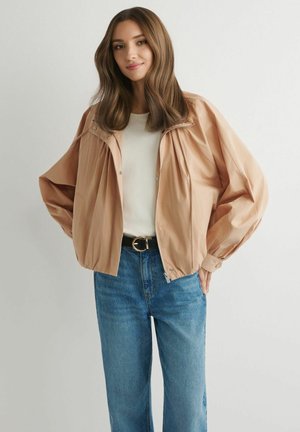 Młoda kobieta o długich brązowych włosach, ubrana w beżową oversize'ową kurtkę, biały top, czarny pasek i niebieskie dżinsy, stojąca na tle jednolitego tła.