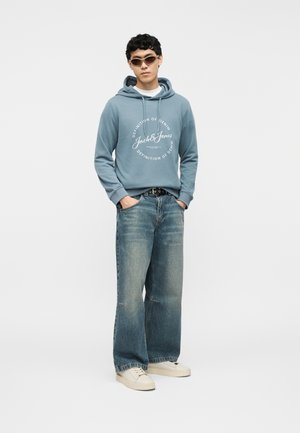 Young man wearing brown sunglasses, blue hoodie with white text, wide-leg faded jeans, and white sneakers, standing with hands in pockets.