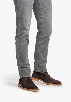 Piernas vestidas con jeans ajustados grises y botas de ante marrón oscuro con cordones hasta el tobillo, con suelas de goma marrón claro.
