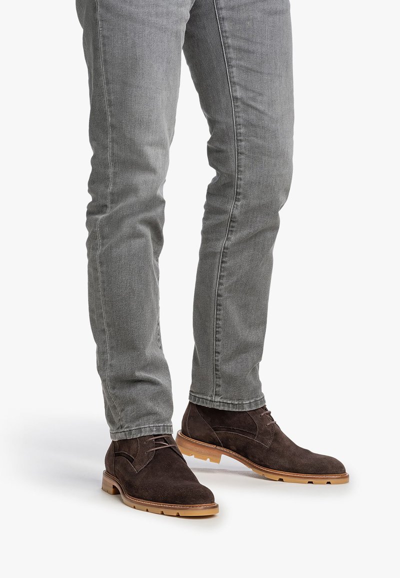 Gambe che indossano jeans slim-fit grigi e stivali alla caviglia in suede marrone scuro con suole in gomma marrone chiaro.