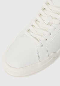 White leather sneaker with flat laces, rounded toe box, and a textured rubber sole. Minimalist design features subtle stitching accents.