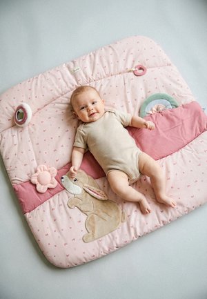 Smiling baby in beige onesie lying on pink play mat with bunny applique, flower toy, and circular mirror on light floor.