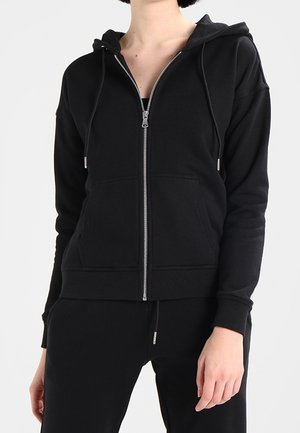 Femme portant un sweat à capuche noir zippé avec des poches avant et un pantalon noir assorti sur un fond uni.