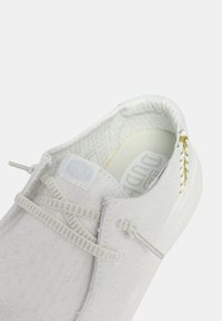 Zapatilla blanca tipo slip-on hecha de tejido texturizado, con un patrón sutil, cordones elásticos y un acento dorado cerca del talón.