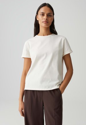 Frau mit langen dunklen Haaren, die ein einfaches weißes kurzärmeliges T-Shirt und dunkelbraune Hosen trägt, stehend vor einem hellgrauen Hintergrund.