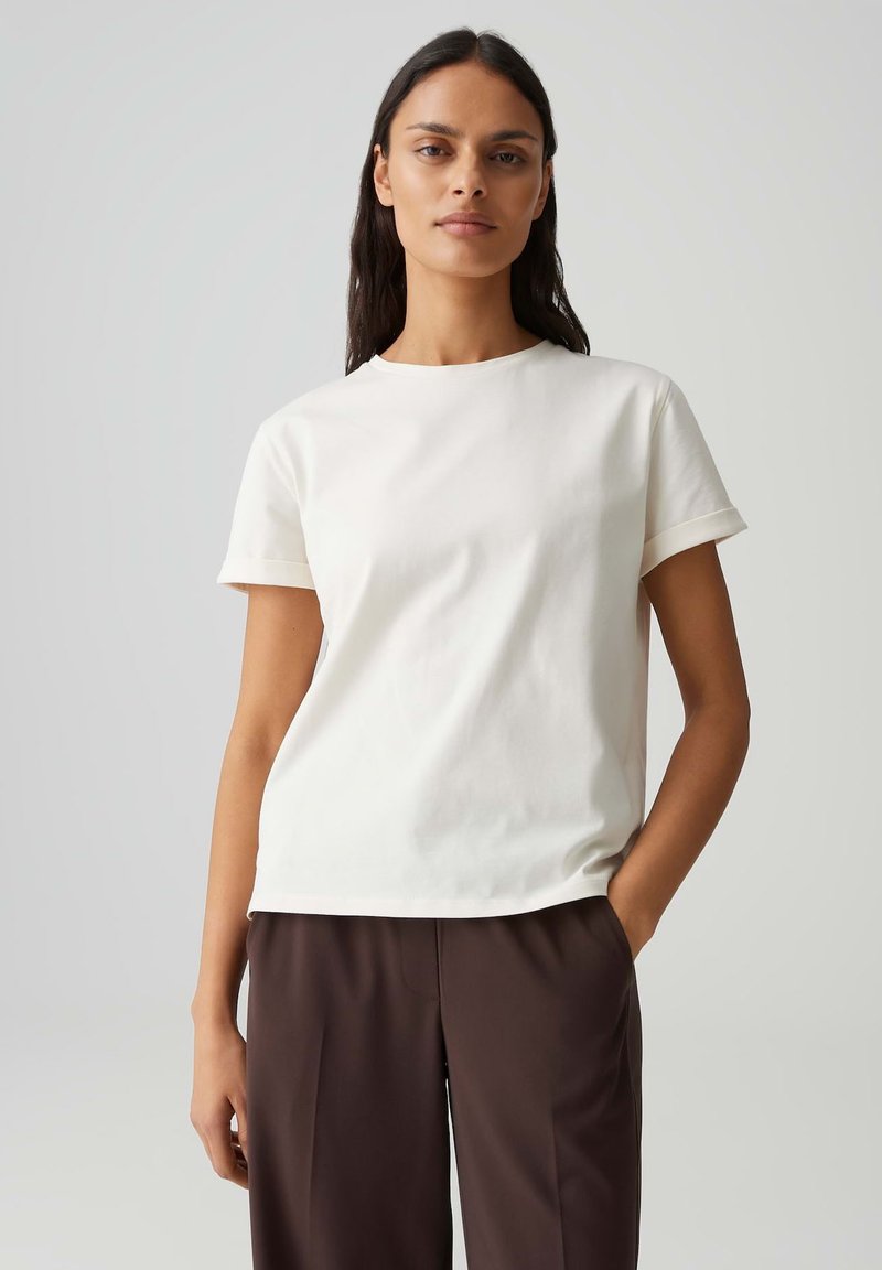 Femme aux longs cheveux noirs portant un t-shirt blanc à manches courtes uni et un pantalon marron foncé, debout devant un fond gris clair.