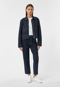 Giacca di jeans scura con cuciture chiare, abbinata a pantaloni di jeans scuri. T-shirt bianca sotto e sneakers bianche. Vestibilità casual, design strutturato.