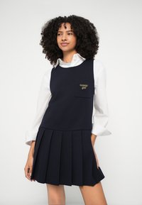 Ναυτικό φόρεμα με πιέτες και λαιμόκοψη σε σχήμα κύκλου, με μπροστινή τσέπη που φέρει το λογότυπο "tommy girl" με χρυσά γράμματα. Φορεμένο πάνω από ένα λευκό πουκάμισο.