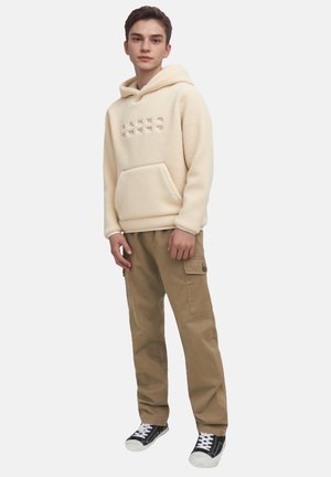 Felpa in pile crema con tasca frontale e testo in rilievo; abbinata a pantaloni cargo beige e sneakers nere con suola bianca.