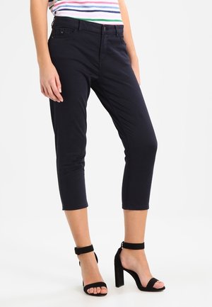Femme portant un pantalon noir court, des sandales à talons hauts avec bride de cheville noires et un haut rayé multicolore.