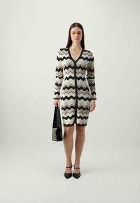 Abito lavorato a maglia con scollatura a V e maniche lunghe, caratterizzato da un motivo a zigzag in nero, bianco e beige. Modella con una borsa nera.