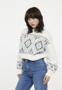 Pull en tricot blanc avec des motifs géométriques bleus et rouges sur la poitrine et les manches, associé à un jean bleu taille haute.