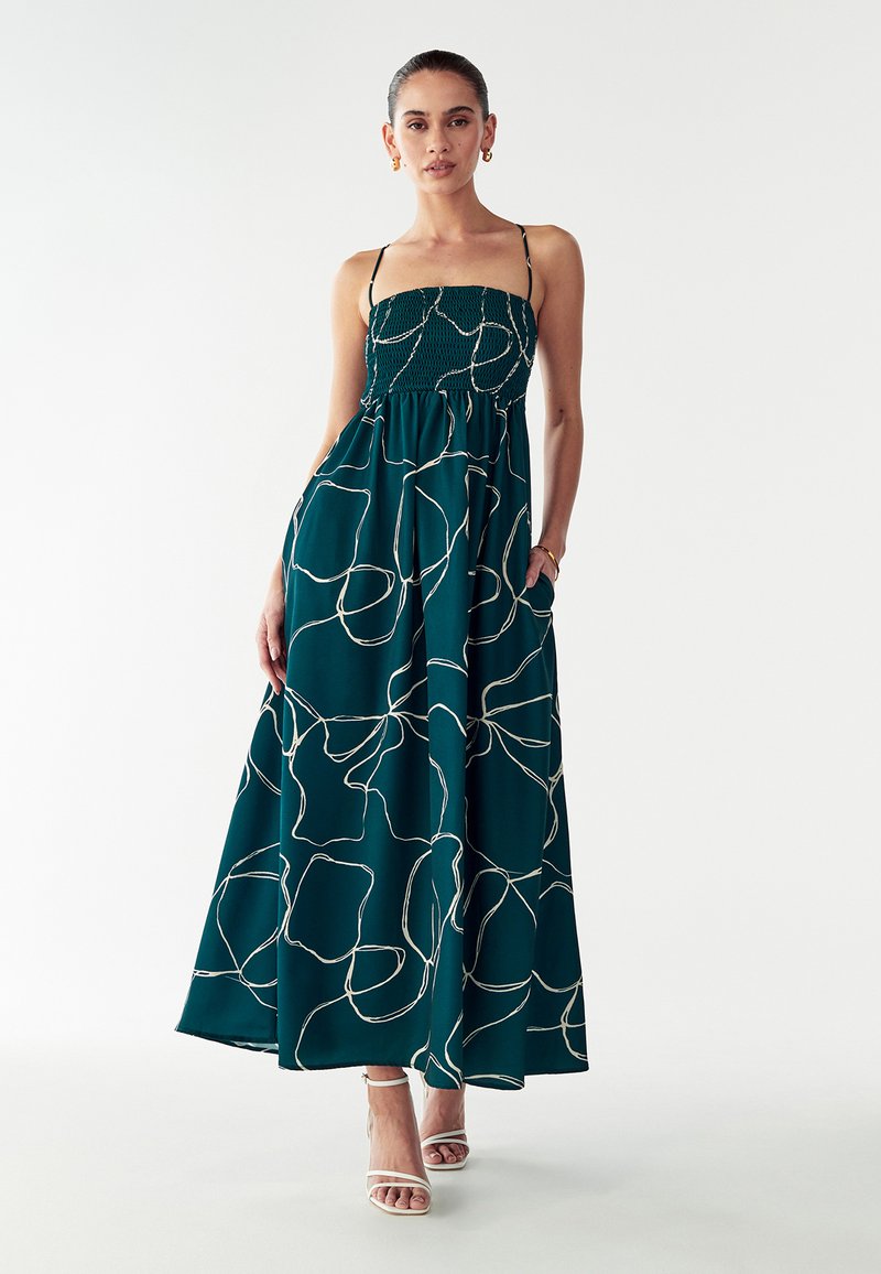 Robe maxi en tissu bleu profond avec des lignes abstraites blanches. Comprend un corsage smocké, des bretelles fines et réglables, et une jupe fluide.