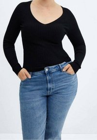Frau trägt ein schwarzes, langärmeliges, geripptes Oberteil mit V-Ausschnitt und blaue hochgeschnittene Jeans, steht mit Händen in den Taschen.