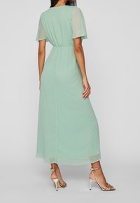 Vrouw in een lichtgroene midi-jurk met korte doorschijnende mouwen en gouden hiëlaar sandalen met bandjes, staand tegen een effen achtergrond.