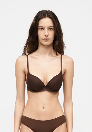 Žena s dugom smeđom kosom, odjevena u smeđi grudnjak i odgovarajući donji rublje, stoji nasuprot obične svijetlosive pozadine, okrenuta prema naprijed.